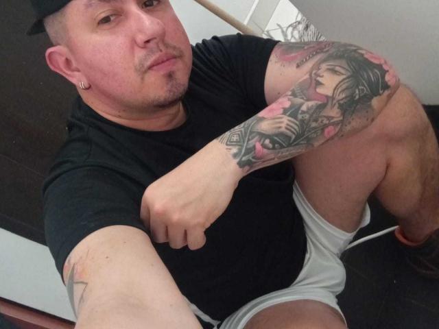 CarlosT69, portant une casquette noire, un t-shirt noir, un short blanc, et avec un bras tatoué représentant une femme avec des roses roses. Il est assis, avec une expression légèrement agacée, et son bras est levé..