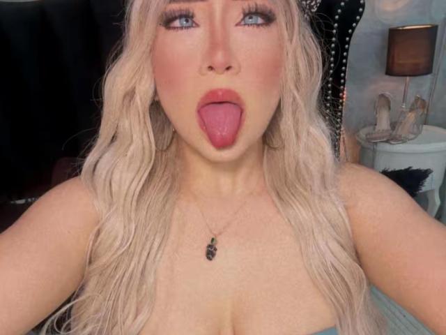 VictoriiaGrey has long, wavy blonde hair and is staring up with blue eyes, her tongue fully out. She has heavy makeup with pink eyeshadow, thick black eyeliner, and red lipstick. She wears a necklace with a small black pendant. Her skin is fair, and she has a noticeable cleavage. The image is a close-up selfie. The background is blurry, but a black curtain and a white table with shoes are visible. The photo is brightly lit.