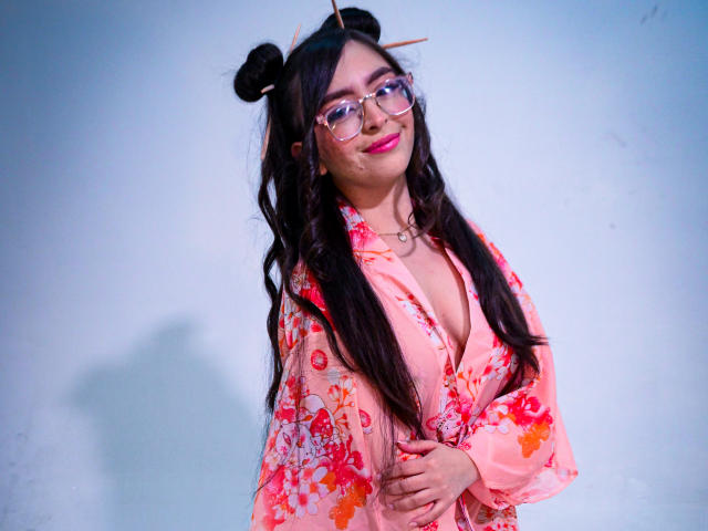 EvaMcAllister with long black hair in buns, glasses, pink floral shirt, heart necklace, smiling, hands on chest, pink lipstick, white wall, soft blue lighting.