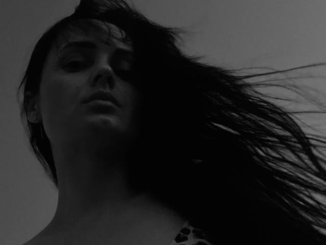 CarolinePirs has dark, flowing hair obscuring her face, with a serious expression. Black-and-white image, low-angle, highlighting her chin and neck. Her tits are partially visible.