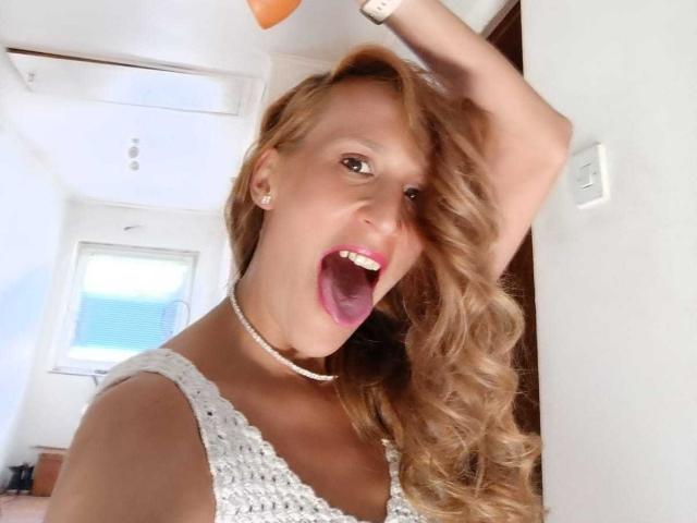 Madame Hermosa, blonde avec des cheveux bouclés, portant un haut blanc en crochet, a la bouche grande ouverte, exprimant la surprise ou l'excitation, et tient son bras levé..