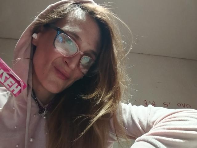MadamHermosa, con lunghi capelli castani, occhiali e una felpa rosa, ha un leggero sorriso ed è china, toccandosi la testa con una mano. Il suo seno è piccolo e la sua vulva non è visibile..