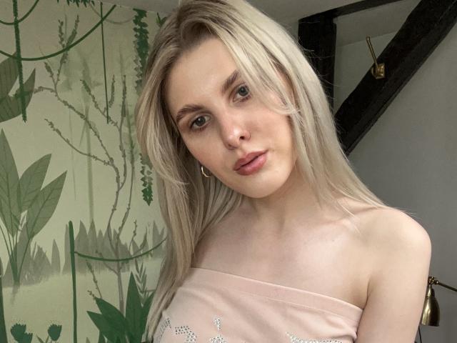 A close-up photo of PinkyZora, a blonde woman with fair skin and blue eyes. She has long, straight hair and is wearing a pink strapless top with small silver embellishments. She has a slight, neutral expression. Her eyebrows are thin and she's wearing small gold hoop earrings. The background has green leafy patterns on a wall, and a black wooden beam is visible on the right. The lighting is natural and even.