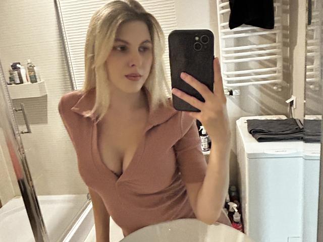 PinkyZora, a blonde woman with shoulder-length hair, takes a selfie in a bathroom. She wears a pink, short-sleeved, V-neck top that reveals moderate cleavage. She holds a black smartphone in her right hand. The bathroom has a white towel radiator on the right, a white sink on the right, and a bathtub on the left with toiletries on a shelf. The photo is slightly blurry.