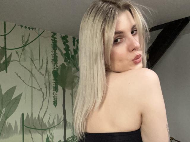 PinkyZora with blonde hair, pouty lips, looking over shoulder, black strapless top, pale skin, green leafy patterned background, black ceiling beam, subtle makeup, slightly messy hair.