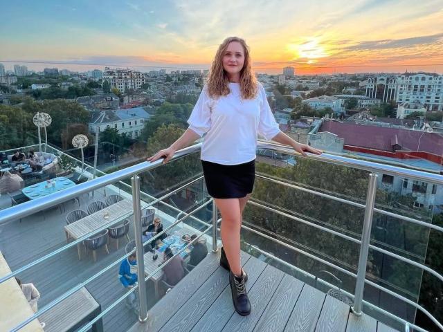 Mirrela with wavy blonde hair, white shirt, black skirt, and black boots, standing on a rooftop, hands on metal railing, sunset in background, city skyline visible.