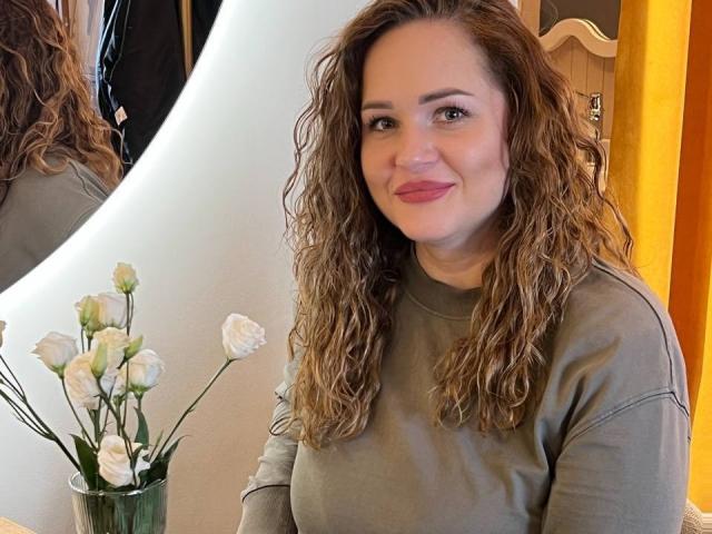 Mirrela with curly brown hair, wearing a gray long-sleeve shirt, sitting, smiling, pink lips, small tits, white roses in a green vase, yellow wall, mirror reflection.