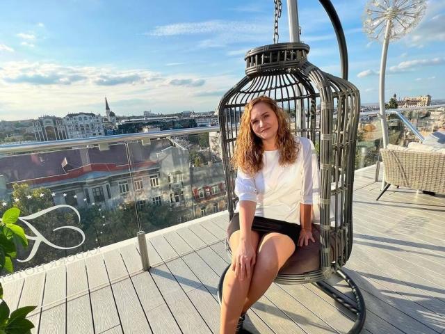 Mirrela sits in a black metal birdcage chair, wearing a white blouse and black skirt, smiling with curly red hair, showing her ass and pussy in the sunlight.
