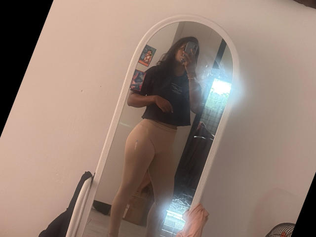 Secretlysweet-hot taking a mirror selfie, wearing a black t-shirt and beige pantyhose, showing her ass, with long dark hair and a mask, in a white-walled room.