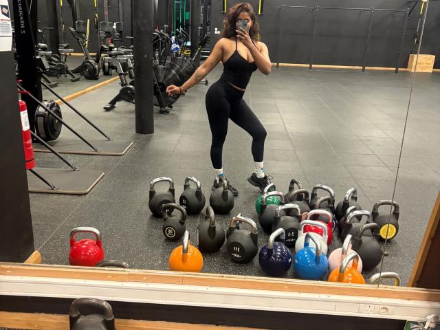 A gym mirror selfie of Secretlysweet-hot, a fit woman with dark curly hair, wearing a black sports bra and black leggings. She stands with one leg raised, holding a smartphone. Surrounding her are multiple kettlebells in black, red, orange, blue, and green, scattered on a grey tiled floor. The gym is equipped with various exercise machines. The image has a casual, real-life photograph quality.