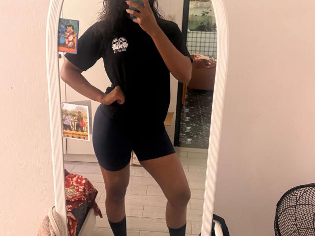 Mirror selfie of Secretlysweet-hot, dark-skinned, wearing black t-shirt, tight blue shorts, socks, standing with hand on hip, reflecting room with bed and wall art.