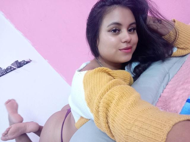 MissyPhil with long black hair, yellow and white sweater, purple thong, lying on a gray couch, smiling, showing her ass, pink wall, and a city skyline poster in the background.