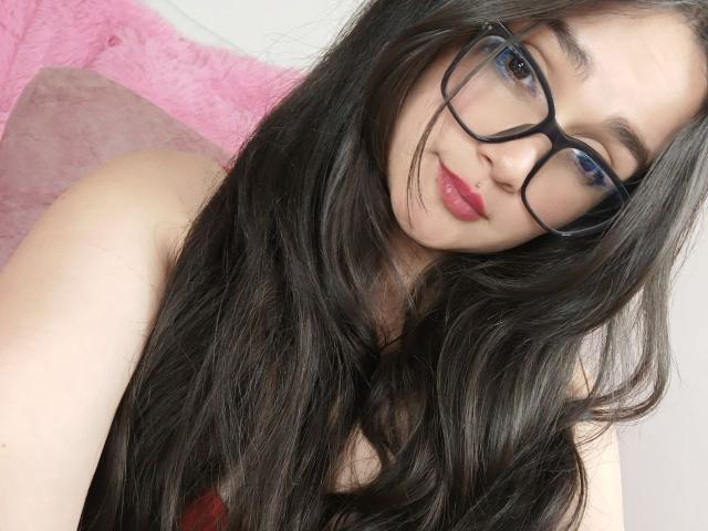 NataliaRivas with long, wavy black hair, wearing black glasses and pink lipstick, looking at the camera with a slight smile, showing her bare shoulders.
