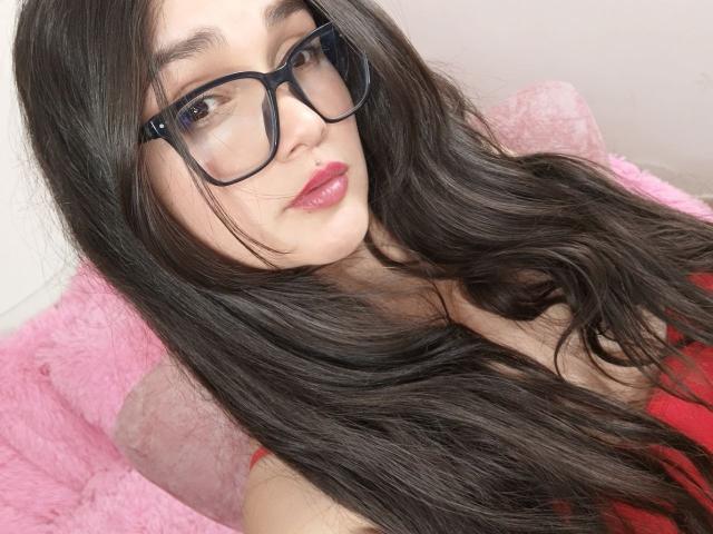 NataliaRivas with long, wavy black hair, wearing red lipstick and black glasses, sitting on a pink fluffy chair, looking at the camera with a slight smile. Boobs visible.
