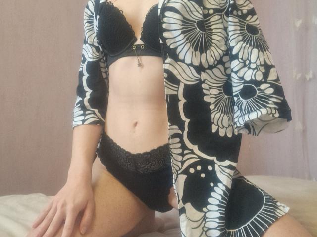 Amberia indossa un reggiseno e una biancheria intima in pizzo nero, con un vestaglie a motivi floreali, che mostrano i suoi seni e i suoi genitali. È inginocchiata su un letto, con le mani sulle cosce..