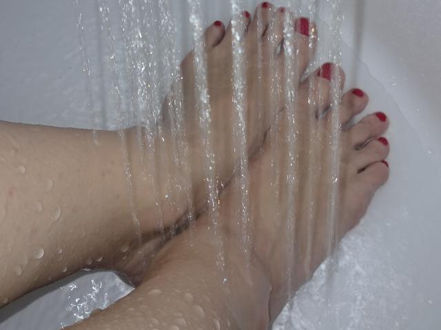 Amberia's feet with red-painted toenails under a stream of water, droplets running down her soles. Her legs are slightly parted, showing her toes.
