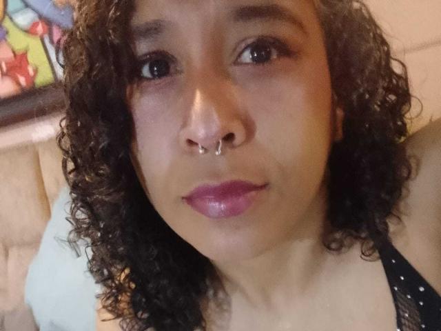 EvelynMonrou with curly brown hair, nose ring, purple lipstick, black lace top, and brown skin, looking at camera with a slight smile. Boobs visible under lace top.