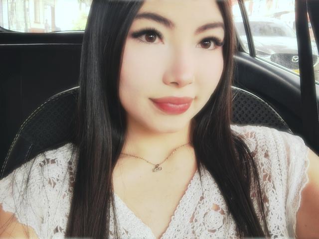 ViolettaLeroy with long black hair, wearing a white lace top, has cute brown eyes, full pink lips, and a small necklace, sitting in a car. Boobs not visible.