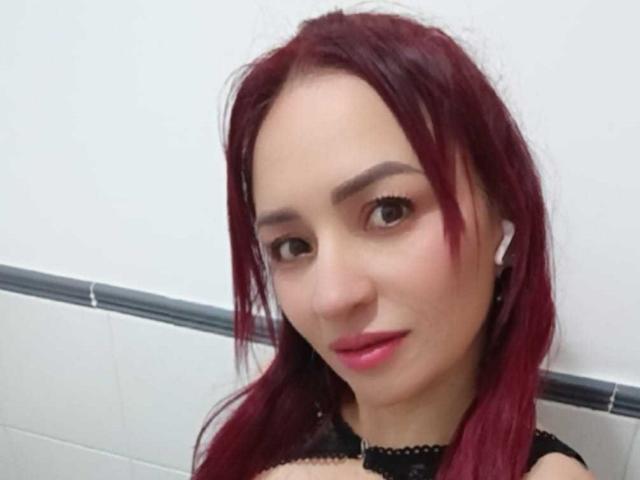 EmaCollins, con i capelli rossi, la pelle chiara e il rossetto rosa, indossa auricolari bianchi e una collana nera. Ha un seno piccolo e un sorriso carino..