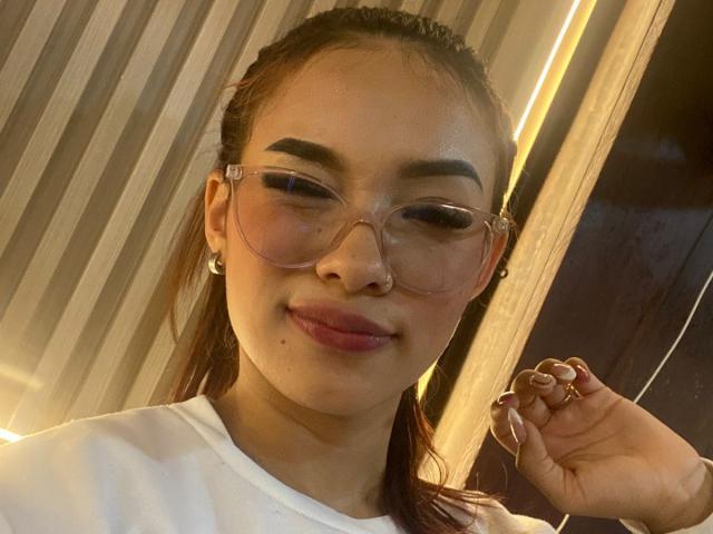SashaHans with brown hair in a ponytail, wearing pink glasses, white shirt, and earrings, smiling with pink lips, holding fingers with pink nails, has a small mole on her cheek.