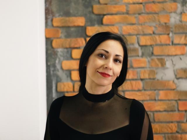 LiluGrants has medium-length black hair, red lipstick, and is wearing a black sheer top with a high neckline. Her tits are covered by the sheer fabric.