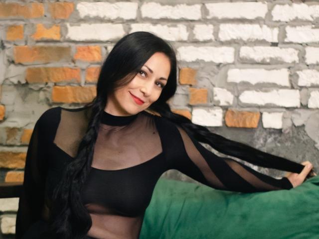 LiluGrants with long black braids, smiling, wearing a sheer black top, revealing her tits, sitting on a green couch, against a brick wall.