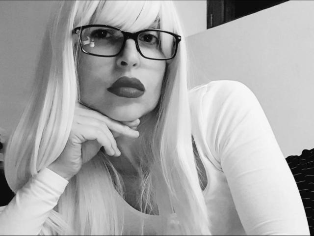 NattyNat, con lunghi capelli biondi, occhiali neri e rossetto scuro, appoggia il mento sulla mano, indossando una maglia bianca a maniche lunghe, in un selfie in bianco e nero..