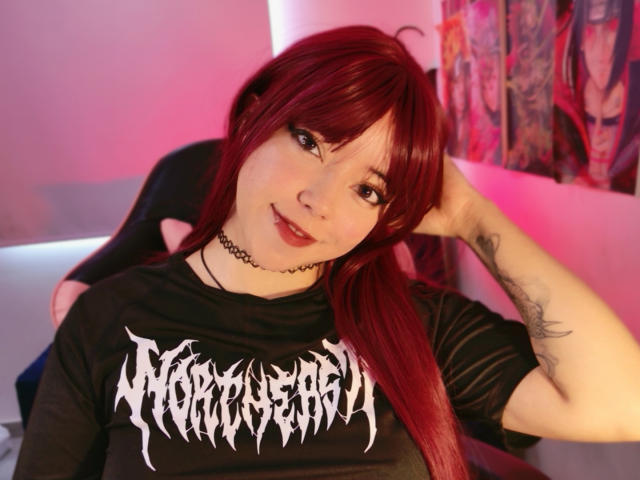 Arieyaara has long, straight red hair with bangs, wearing a black t-shirt with white, Gothic-style text. She has light skin, a black choker necklace, and a tattoo on her left forearm. She's looking at the camera with a slight smile, her right hand touching her hair. The image has a pink and purple light, with colorful, abstract-patterned curtains in the background.