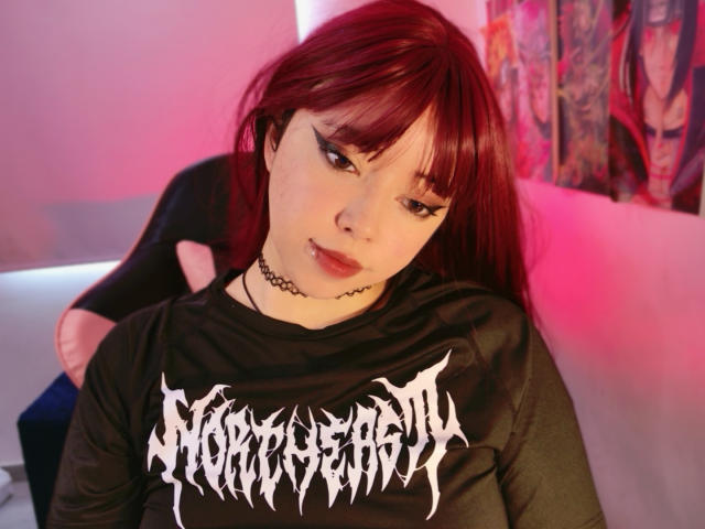 Arieyaara has straight, dark red hair with bangs and wears a black, long-sleeve shirt with white, gothic-style text on the chest. She has pale skin, dark eyeliner, and a black choker. Arieyaara is sitting with her back to the camera, looking slightly down and to the left. The room has pink and purple lighting. The background shows a colorful, abstract pattern on the right. The image is focused on Arieyaara's face and upper body.