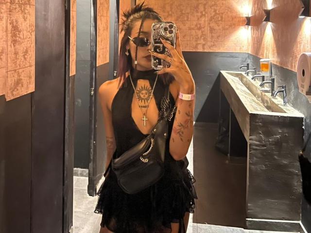 SalomeMille with tattoos, black halter top, deep cleavage, black skirt, black cross necklace, black handbag, holding phone, in bathroom mirror selfie. SalomeMille has black hair, sunglasses.