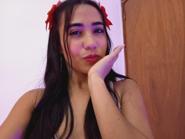 KateWendys with long black hair, red bows, pink eyeshadow, and red lipstick, pouting with her hand on her cheek, wooden door in background, purple lighting on her face.