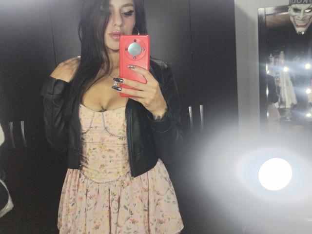 ErotiicAlexa with long brown hair, wearing a black leather jacket, pink floral dress, showing her tits, holding a pink phone, mirror selfie, reflection of bright lights.