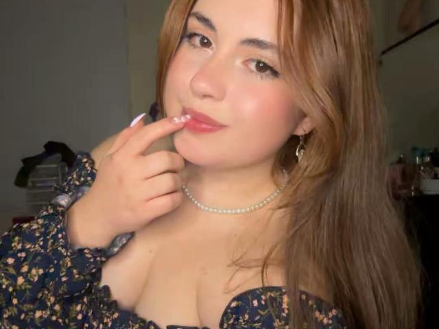 VicluX with light brown hair, wearing a floral dress, pearl necklace, and earrings, touching her pink-lipped lips with her finger, showing her tits and a slight smile.