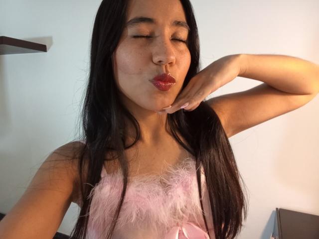 KateWendys, an Asian woman with long black hair, is in a pink, furry, lace-trimmed top. She's puckering her lips, making a kissy face, with her right hand touching her cheek. Her eyes are closed, and she's smiling slightly. The light is even, highlighting her smooth skin and subtle makeup. Her top has a small bow in the center. The background is mostly white with a shelf on the left. The image is casual and focused on her playful expression.