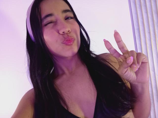 KateWendys with long black hair, pink lips, and a cute pout, winking and making a peace sign with her right hand, wearing a black top, purple light in background.