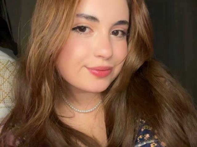 VicluX has light brown wavy hair, a cute face with pink lips, a pearl necklace, and is smiling slightly at the camera with brown eyes and subtle makeup.