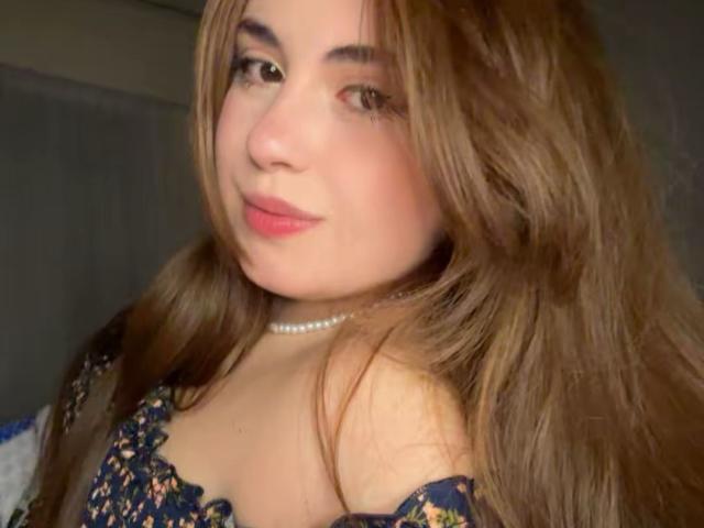 VicluX has long brown hair, a pearl necklace, and a floral off-shoulder top, showing her cute face and small tits in a slightly blurry selfie.