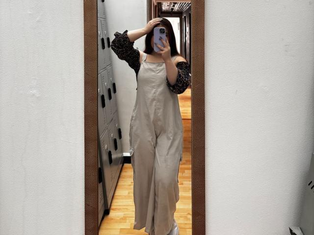VicluX in a beige dress with black polka dot sleeves, holding a phone, covering her head, standing in front of a mirror, wooden floor, white walls, reflection.