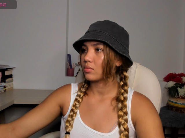 KataSofia with braided blonde hair, wearing a grey bucket hat and white tank top, sitting in a chair, looking slightly to the side with a neutral expression.