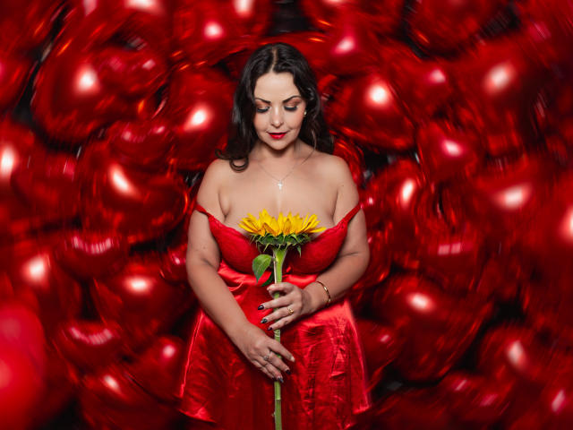 MelissaLeen with big tits, holding a sunflower, wearing a red dress, surrounded by red heart-shaped balloons, smiling with red lipstick, and dark wavy hair.