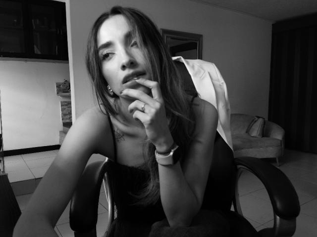 AdharaCooperr with long hair, wearing a black dress, leaning forward in a chair, finger near her slightly open mouth, showing her tits, and a ring on her finger. Black-and-white.