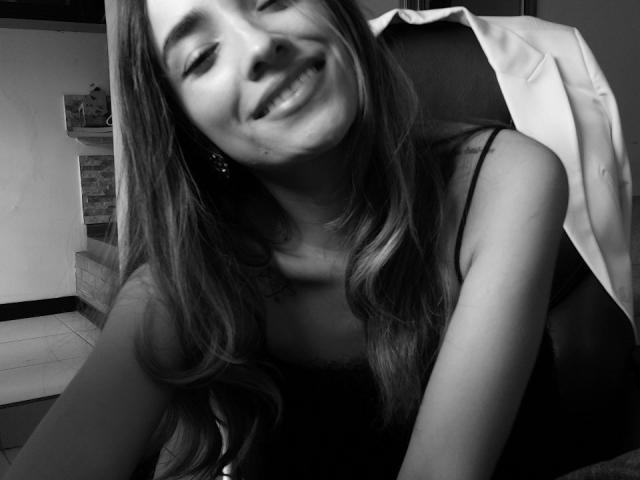 Black and white photo of AdharaCooperr with long wavy hair, smiling, wearing a black dress, and a white jacket draped over a chair, showing her cute, small tits.
