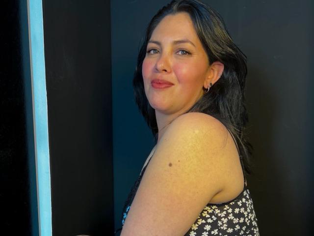 Irisbloom69, a chubby woman with dark hair and fair skin, wearing a floral dress, smiling slightly with pink lips, small earrings, and a small mole on her upper arm.