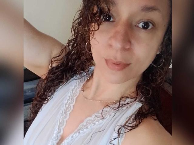 Angelonix with curly brown hair, wearing a white lace-trimmed top, has a slight smile and brown eyes, showing her small tits and a delicate necklace.