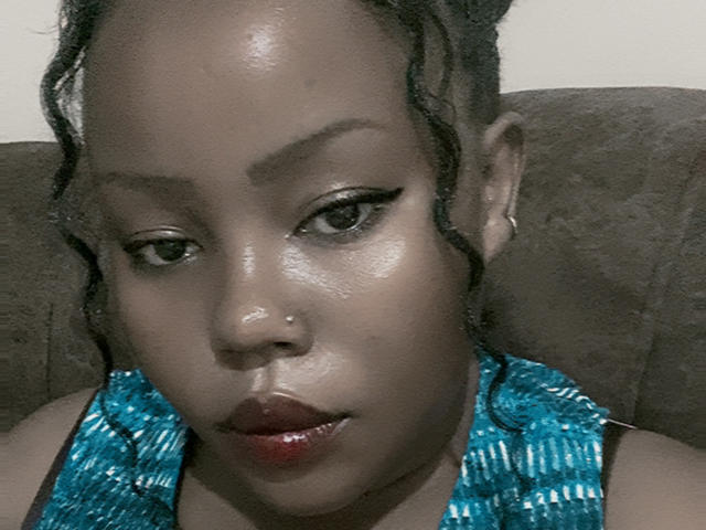 AnitaNini with shiny brown skin, braided hair, winged eyeliner, red lipstick, blue shiny top, nose piercing, and small hoop earring, sitting on a brown sofa.