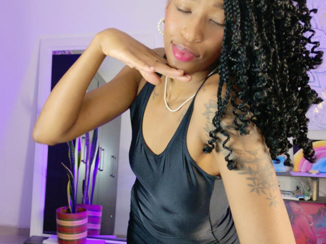 ImSelma with dark skin, curly black hair, and a tattoo on her right arm, wearing a black dress, touching her chin with a playful expression and pink lipstick.