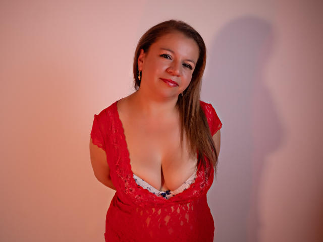 RosieRider with brown hair, red lace top, deep cleavage, showing her big boobs, smiling, hands behind back, pink shadow on wall, wearing red earrings.