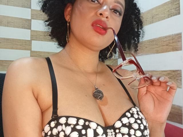 VioletaSwitch with curly black hair, red lipstick, holding red glasses, black strap bra with white patterns, showing her big tits, wearing gold earrings and a heart necklace.