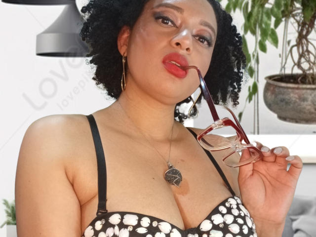 VioletaSwitch has a medium-brown complexion and curly black hair. She wears red lipstick, gold hoop earrings, and a black bra with white floral patterns. She holds red glasses in her right hand, which are near her slightly open mouth. She has a heart-shaped pendant on a thin necklace. The background includes a potted plant and a grey lamp. The image has a watermark with the text "Love love love."