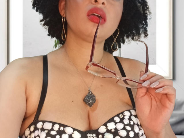 VioletaSwitch with curly black hair, holding pink glasses in her mouth, wearing a black and white polka dot bra, gold hoop earrings, and a leaf necklace, tongue slightly out.